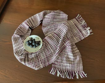 May include: A white and purple patterned scarf with fringe on the ends. The scarf is folded in a loop and a small white plate with purple grapes is sitting on top of the loop.