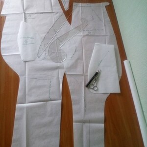 May include: A set of white paper sewing patterns for pants, with markings and instructions. The patterns are laid out on a wooden surface.