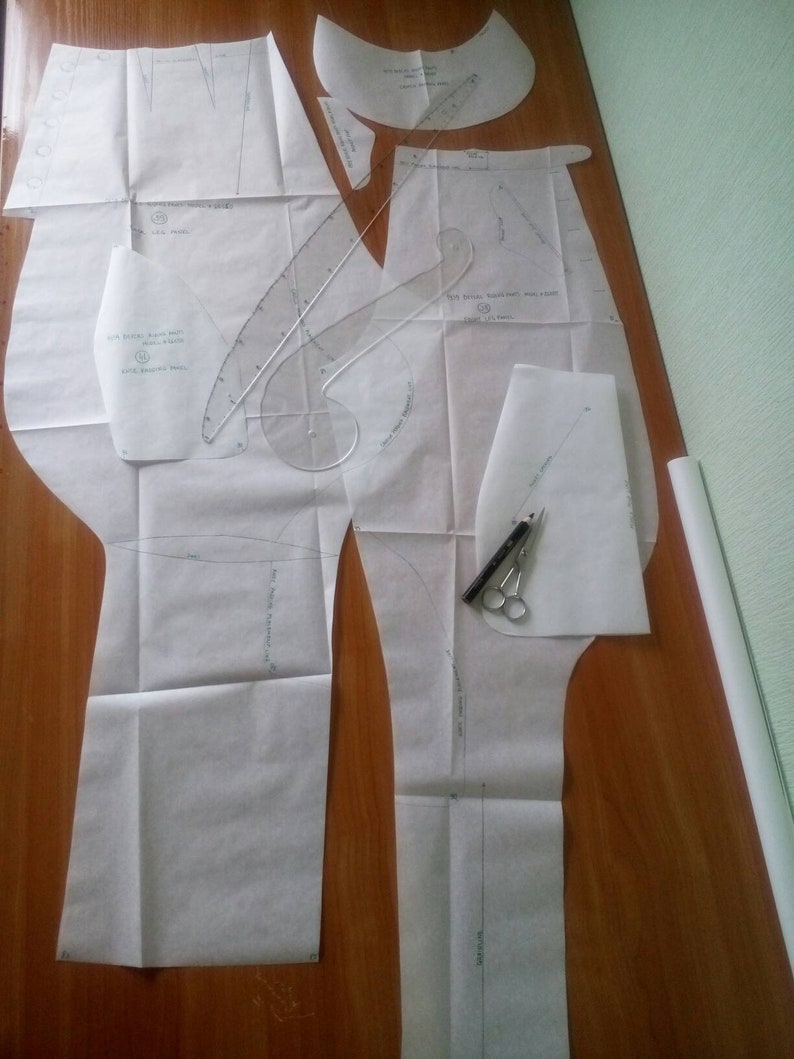 May include: A set of white paper sewing patterns for pants, with markings and instructions. The patterns are laid out on a wooden surface.