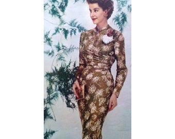 May include: A woman wearing a brown and white floral dress with a white flower brooch. She is holding a small gold clutch.