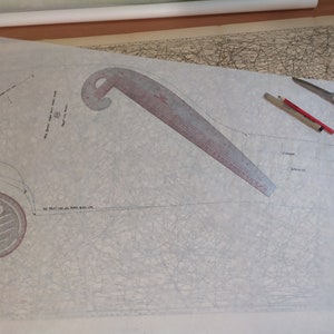 May include: A white sheet of paper with a red and white curved ruler, a red pencil, and a pair of black scissors. The paper has a grid pattern and some handwritten text.