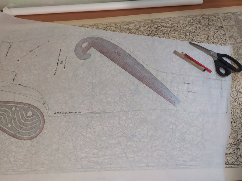 May include: A white sheet of paper with a red and white curved ruler, a red pencil, and a pair of black scissors. The paper has a grid pattern and some handwritten text.