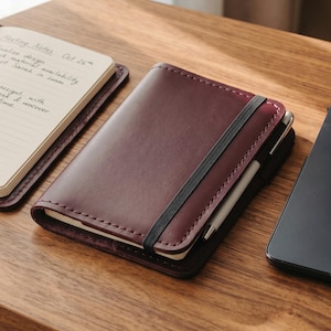 Op de afbeelding: Een bordeauxrood leren notitieboek met een zwarte elastische band en een pen, naast een open notitieboek met handgeschreven notities. De tekst "Meeting Notes Oct 26" is zichtbaar. De items staan op een houten oppervlak, met een laptop op de achtergrond.