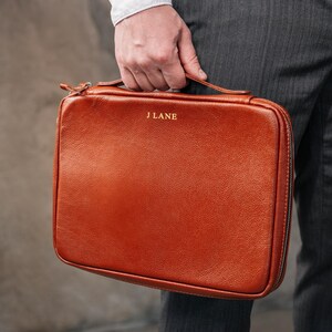 May include: A rectangular, brown leather briefcase with a top handle and a wraparound zip. The text "J LANE" is embossed in gold on the front. The briefcase is held by a person wearing a white shirt and grey trousers.
