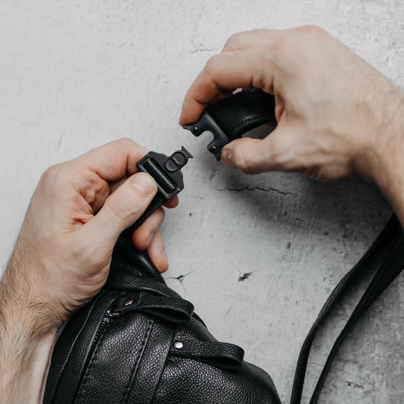 Puede incluir: Bolso bandolera de cuero negro con superficie texturizada. El bolso tiene un cierre de hebilla negro. La imagen muestra la hebilla que se est&aacute; abrochando. El bolso tiene una cremallera y una correa.