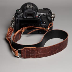 May include: A brown leather camera strap with a crocodile texture, attached to a black DSLR camera. The strap has a black underside and silver metal rings for attachment. The camera is black.
