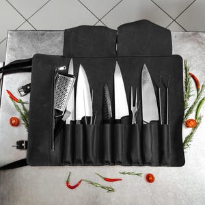 Könnte beinhalten: Eine schwarze Messertasche aus Leder mit verschiedenen Küchenwerkzeugen aus Edelstahl, darunter mehrere Kochmesser, eine Fleischgabel, eine Reibe und Metallzangen, die in einzelnen vertikalen Fächern mit Klappdeckel und Riemen verstaut sind.