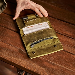 May include: An olive green leather tobacco pouch featuring a dedicated slot for a lighter, a central compartment for rolling papers, and a zippered pocket. The wallet-style accessory is constructed with visible stitching and two metal snap fasteners.