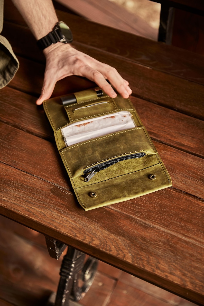 May include: An olive green leather tobacco pouch featuring a dedicated slot for a lighter, a central compartment for rolling papers, and a zippered pocket. The wallet-style accessory is constructed with visible stitching and two metal snap fasteners.