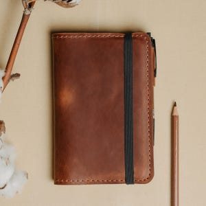 Op de afbeelding: Een bruin lederen notitieboek met zichtbare stiksels langs de randen, een verticale zwarte elastische bandsluiting en een penhouder aan de rechterkant. Het notitieboek ligt naast een bruin houten potlood en een gedroogde katoentak.