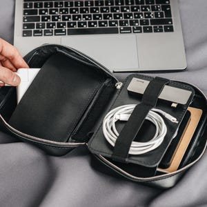Puede incluir: Un organizador tecnológico de cuero negro con cremallera, abierto para revelar un cargador blanco, un cable blanco, una unidad USB plateada y un teléfono. Un portátil y un smartphone están en el fondo.