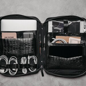 May include: A black zipped organiser case with a crocodile-embossed texture, featuring multiple interior compartments, slots for pens, and elastic loops holding white charging cables. It contains a laptop, a notebook labelled THE FUTURE, and a tablet.