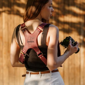 Può includere: Un'imbracatura per fotocamera in pelle bordeaux con elementi metallici argentati. L'imbracatura presenta un design centrale con la scritta "PHOTOGRAPHER" e la silhouette di una persona che tiene una fotocamera. La persona indossa un top nero e pantaloni bianchi.