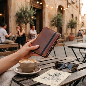 May include: A brown leather notebook with a black elastic band closure and a silver pen attached to the side. The notebook features the embossed letters O.W. on the front cover. It is held in a hand near a ceramic coffee cup and sunglasses.