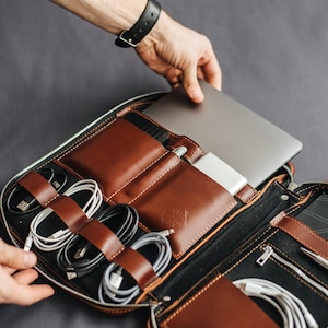 Può includere: Un organizer tecnologico in pelle marrone con più scomparti, tra cui una custodia per laptop, un portapenne e organizzatori di cavi. La custodia è aperta, rivelando un laptop, cavi e accessori. L'organizer è maneggiato da una persona.