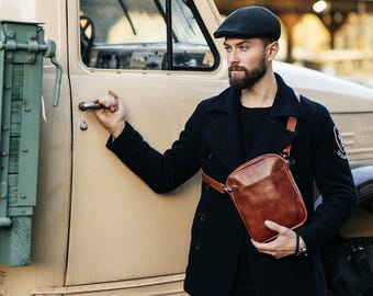 May include: A brown leather crossbody bag with a shoulder strap. The bag is rectangular with a flap closure. The person is wearing a black coat and a dark cap. The background includes a vintage truck.