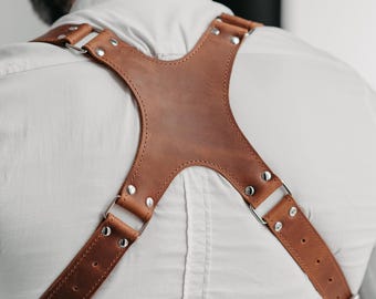 May include: A brown leather harness featuring a central X-shaped back panel, adjustable straps with multiple punched holes, silver-toned metal rivets, and rectangular buckles. The leather has a smooth, matte finish with visible stitching along the edges.