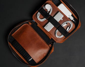 May include: A brown leather tech organizer case with a zipper closure. The interior features a large open pocket and a section with black elastic straps holding white charging cables, a power adapter, and a portable power bank device.