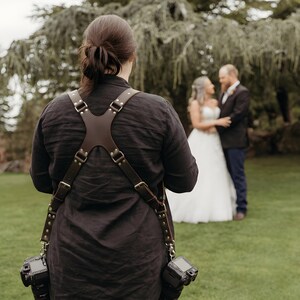 May include: A dark brown leather camera harness with an X-shaped back panel, adjustable straps, and metal buckles. The harness features two metal clips at the bottom, each attached to a black digital camera, worn over a dark long-sleeved shirt.