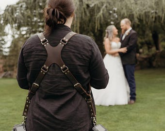 May include: A dark brown leather camera harness with an X-shaped back panel, adjustable straps, and metal buckles. The harness features two metal clips at the bottom, each attached to a black digital camera, worn over a dark long-sleeved shirt.
