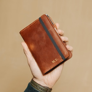 Peut inclure: Un portefeuille en cuir marron doté d'une bande élastique noire verticale sur la couverture avant. Le coin inférieur droit du portefeuille est gravé des initiales M.T. en couleur dorée. Le portefeuille présente une finition mate et lisse.