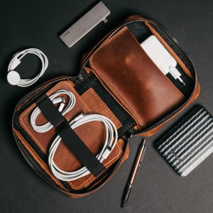 Puede incluir: Un organizador de viaje de cuero marrón con cremallera, que contiene un cargador blanco, cables y un bolígrafo. Un concentrador gris, un cargador de Apple Watch blanco y un banco de energía a rayas también son visibles sobre una superficie negra.
