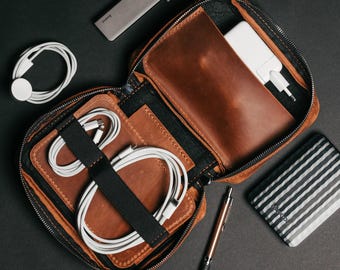 Könnte beinhalten: Ein brauner Leder-Reiseorganizer mit Reißverschluss, der ein weißes Ladegerät, Kabel und einen Stift enthält. Ein grauer Hub, ein weißes Apple Watch-Ladegerät und eine gestreifte Powerbank sind ebenfalls auf einer schwarzen Oberfläche zu sehen.