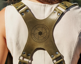 May include: An olive green leather camera harness featuring a central X-shaped back plate with silver-tone metal rivets and hardware. The back plate is embossed with a circular logo containing a camera aperture icon and the text J.C. LUCATERO PHOTOGRAPHY 20 24.