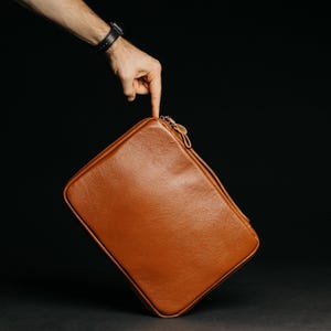 May include: A brown leather case with a zip closure. The case is rectangular with rounded corners. The image is set against a black background.