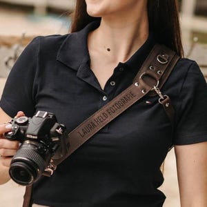 May include: A brown leather camera strap with the text "LAURA HELD FOTOGRAFIE" is draped over the shoulder of a person wearing a black polo shirt. A black Nikon camera is attached to the strap. The strap has metal rings and a clasp.
