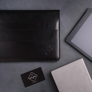 以下が含まれることがあります： フラップ開閉とエンボスロゴが付いた黒い革製タブレットスリーブ。タブレット、小さな灰色のノート、名刺も濃い灰色の表面にあります。名刺にはロゴとテキストがあります。