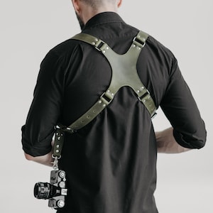 May include: An olive green leather camera harness featuring a cross-back design with silver-toned metal hardware, rivets, and adjustable straps. The harness is worn over a black shirt and supports a black camera hanging from a metal clip.