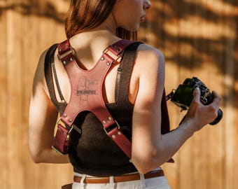 May include: A maroon leather camera harness featuring an X-shaped back panel with a laser-engraved illustration of a photographer and the text @POPLEWSKAPHOTO. The harness is secured with silver-toned metal hardware, including rivets and rectangular rings.