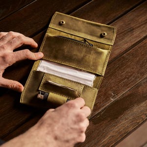 May include: An olive green leather tobacco pouch featuring a zippered pocket, a dedicated slot for rolling papers, and a small loop for a lighter. The interior is organized with multiple compartments and secured with two metal snap fasteners.