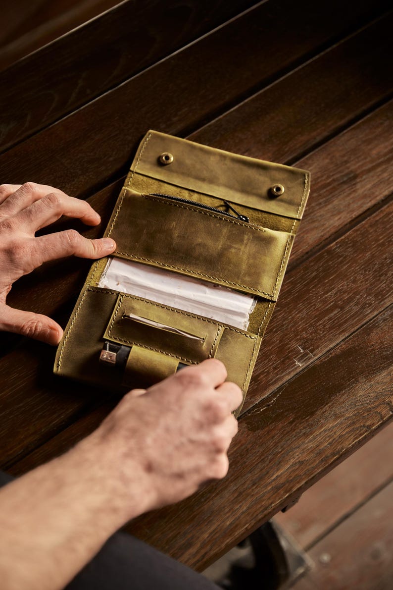 May include: An olive green leather tobacco pouch featuring a zippered pocket, a dedicated slot for rolling papers, and a small loop for a lighter. The interior is organized with multiple compartments and secured with two metal snap fasteners.