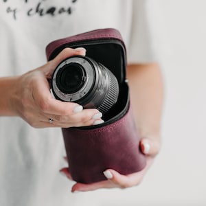 May include: A deep burgundy leather camera lens case with a zippered opening, containing a black camera lens. The case features a structured, upright design with a smooth matte finish, providing a protective storage solution for photography equipment.