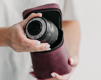 May include: A deep burgundy leather camera lens case with a zippered opening, containing a black camera lens. The case features a structured, upright design with a smooth matte finish, providing a protective storage solution for photography equipment.