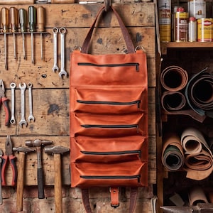 May include: A hanging orange leather tool roll featuring five horizontal zippered compartments, a top carrying handle, and a bottom buckle strap closure. The organizer is suspended from a metal hook against a rustic wooden workshop wall.