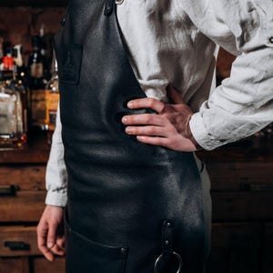 May include: Black leather apron with a large front pocket and a metal ring attached. The apron has a textured surface and adjustable straps. The apron is designed for use in a bar or kitchen setting.