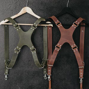 May include: Two leather cross-back suspender harnesses, one in olive green and one in brown. Each harness features adjustable straps, silver-toned metal hardware, rivets, and swivel snap hooks at the ends for attachment, displayed on hangers.