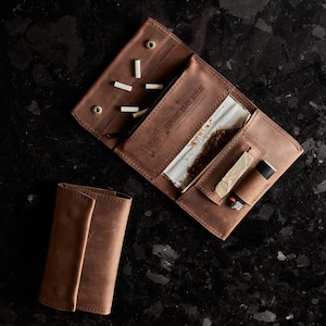 May include: A brown leather tobacco pouch with a flap closure, featuring an open interior with a zippered pocket, a compartment for rolling papers, and a dedicated slot for a lighter. The leather is embossed with the text EXPLORE. ENJOY. REPEAT.