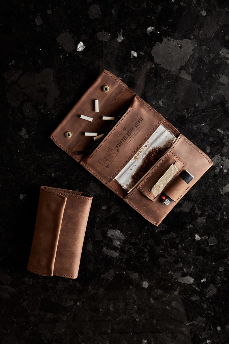 May include: A brown leather tobacco pouch with a flap closure, featuring an open interior with a zippered pocket, a compartment for rolling papers, and a dedicated slot for a lighter. The leather is embossed with the text EXPLORE. ENJOY. REPEAT.