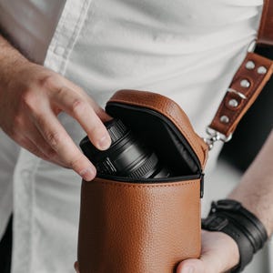 May include: A brown leather camera lens case with a black zip, holding a black camera lens. The case has a matching brown leather strap. The image shows a person holding the case.
