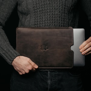 Può includere: Una custodia per laptop in pelle marrone scuro con il logo Apple in rilievo sul davanti. La custodia è tenuta davanti a una persona che indossa un maglione grigio scuro. Il laptop è argentato.