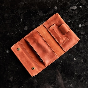 May include: A tan leather wallet featuring a tri-fold design with visible stitching, two brass-colored snap button closures, a central pocket, and a dedicated pen loop on the right side. The interior displays a smooth, matte finish throughout.
