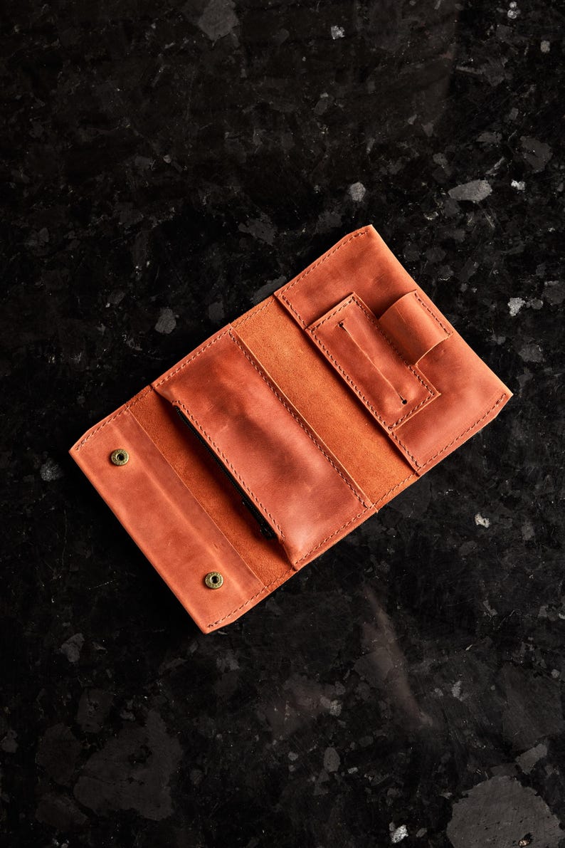 May include: A tan leather wallet featuring a tri-fold design with visible stitching, two brass-colored snap button closures, a central pocket, and a dedicated pen loop on the right side. The interior displays a smooth, matte finish throughout.