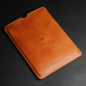 May include: A tan leather tablet sleeve with the Apple logo embossed in the center. The word "DAVID" is subtly printed at the bottom right corner. The sleeve has a stitched border and a cutout at the top.