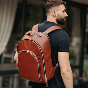 May include: A medium-sized, smooth, cognac-brown backpack featuring a rounded top silhouette, a large front zippered pocket, and a top carry handle. The bag includes silver-toned metal zippers with matching brown leather pull tabs and adjustable straps.