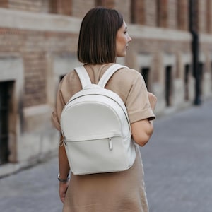 Op de afbeelding: Een witte, afgeronde rugzak met een handvat aan de bovenkant en verstelbare schouderbanden. De rugzak heeft een voorvak met rits en een minimalistisch ontwerp. De tas is gemaakt van een glad materiaal en wordt gedragen.