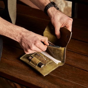 May include: An olive green leather wallet featuring a fold-over design with a snap button closure. The interior includes a zippered compartment, a dedicated slot for a small cylindrical tool, and an open pocket containing white paper documents.